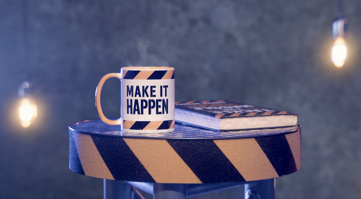 Image of a cup with the logo 'Make it happen' and a book on a work table .